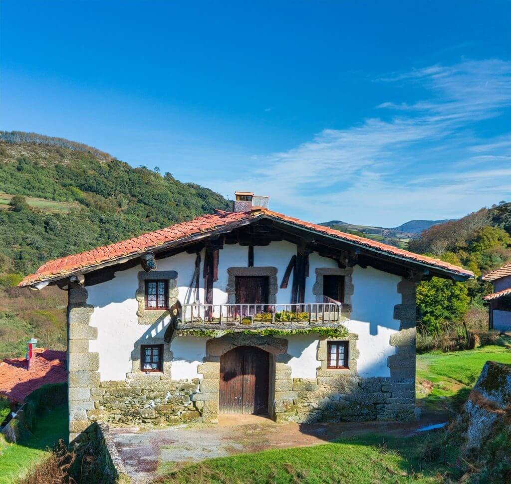 Caserío de Piedra en País Vasco Fotografía de un caserío vasco tradicional, con estructura de piedra y madera, tejado inclinado y grandes ventanas, situado en un entorno rural rodeado de verdes colinas y bosques, representando la arquitectura típica del norte de España.
