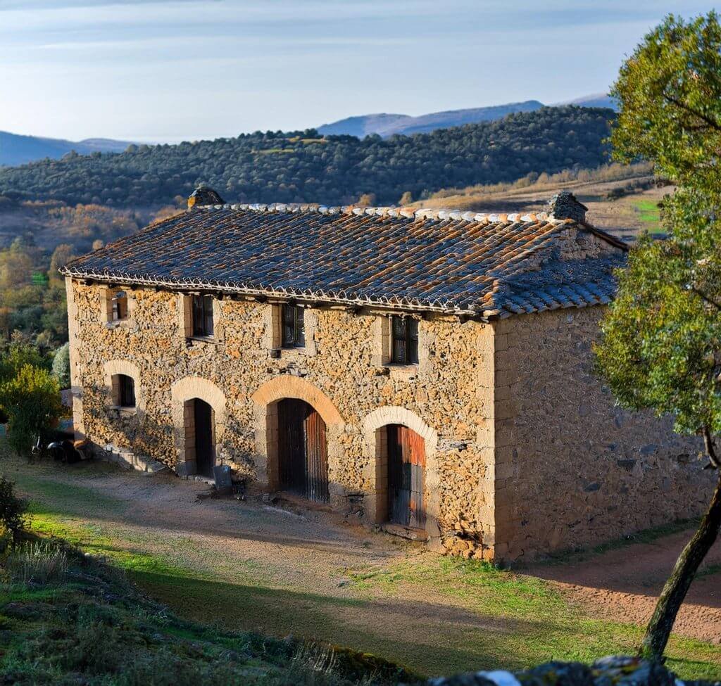 Masía catalana de piedra Fotografía de una masía catalana tradicional en un paisaje rural, con paredes de piedra natural y techo de tejas envejecidas, rodeada de colinas y vegetación, representando el encanto rústico y la arquitectura histórica de las casas rurales del norte de España.
